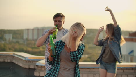 The-American-blonde-woman-is-dancing-on-the-roof-with-her-friends-on-the-party.-She-smiles-and-enjoys-the-time-in-shorts-and-she-is-dancing-with-a-beer-in-a-gray-tank-top-and-a-dark-green-plaid-shirt.